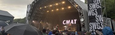 A view of CMAT performing onstage with an audience outside in the rain.