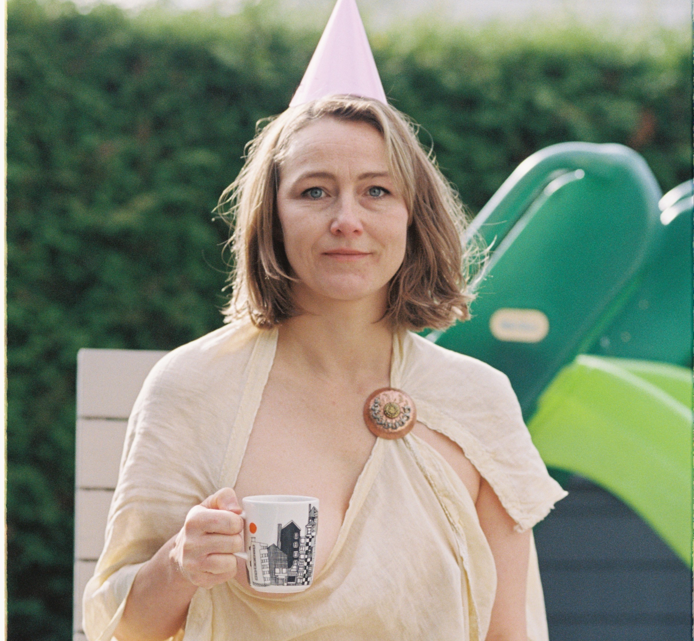 An image of Cathrine Frost in a garden, wearing a cone party hat, with a mug of tea.