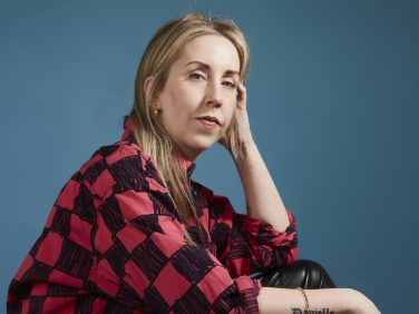 Gemma Rolls-Bentley sat looking at the camera with a blue background. She is wearing a red and black shirt.