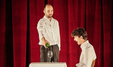 Two actors in white shirts with a red curtain background. One is pouring wine for the other.