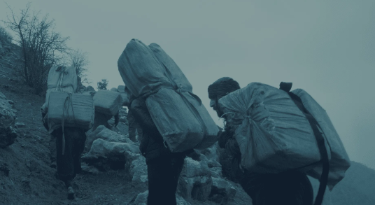 A dark grey image of people walking up a steep mountain with large packs on their backs.