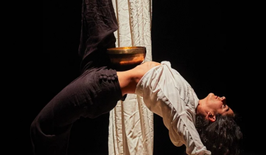 A photograph of the performer, Ashtar Muallem, in a backbend with a golden bowl balanced on their torso.