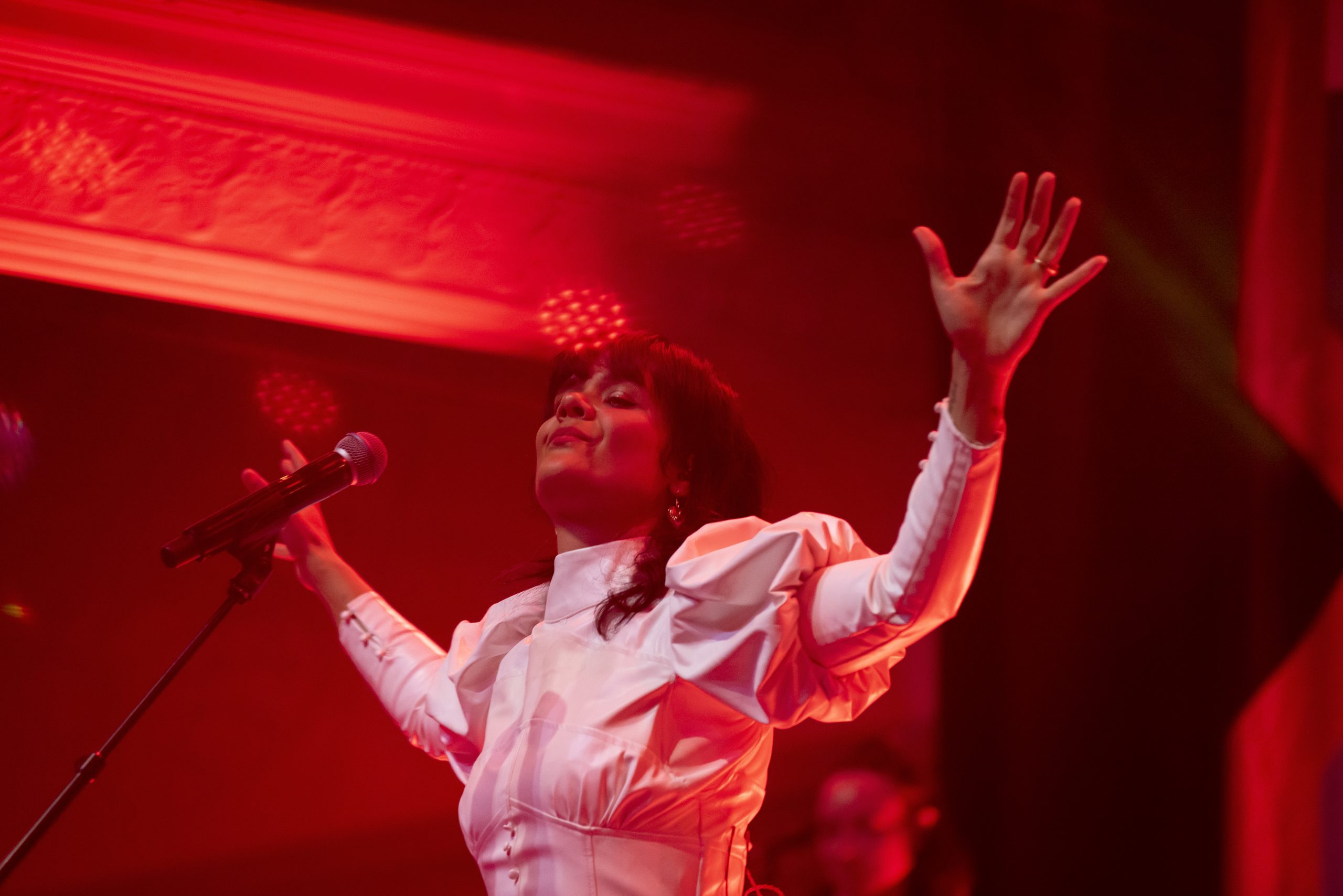 Bat For Lashes performing at The Queen's Hall in Edinburgh in a white dress, bathed in red light, hands raised up.
