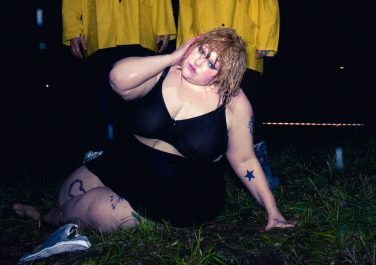 Beth Ditto in a black outfit on the grass in the dark, with two figures behind her dressed in yellow.
