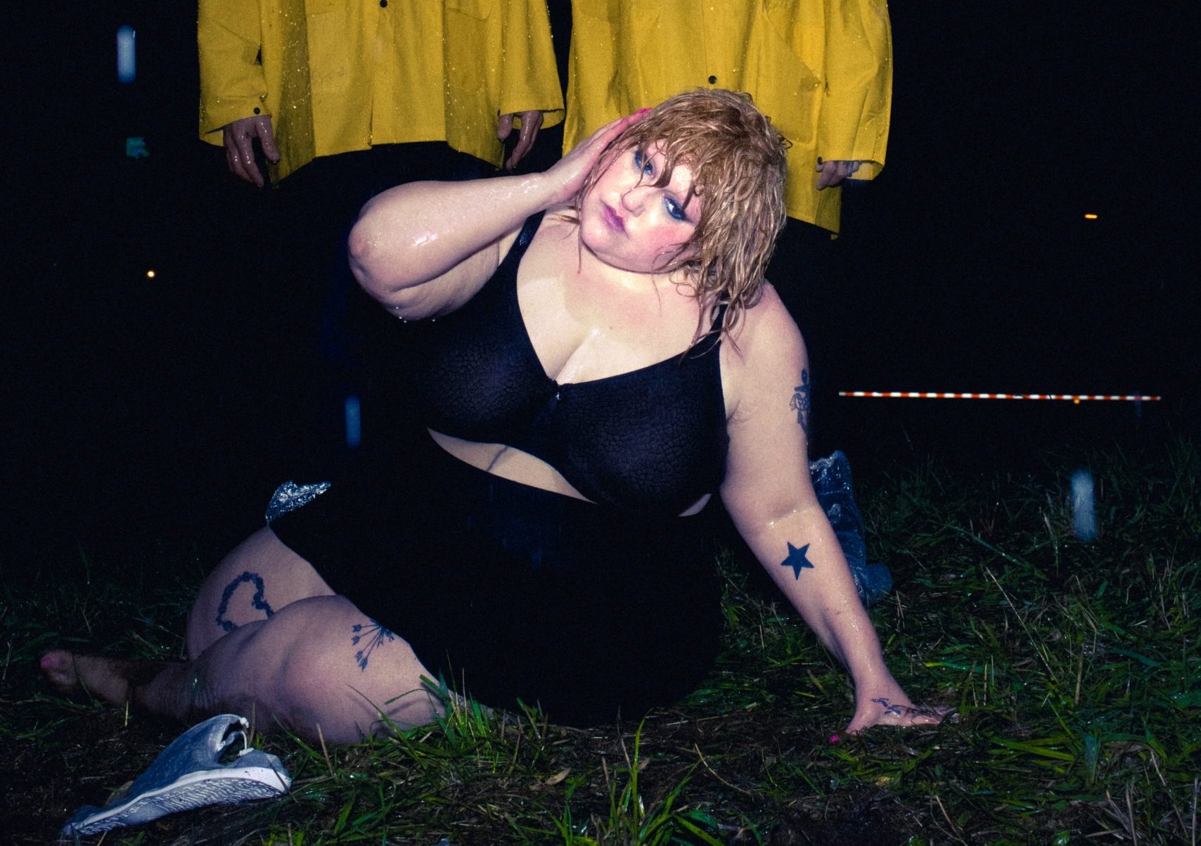 Beth Ditto in a black outfit on the grass in the dark, with two figures behind her dressed in yellow.
