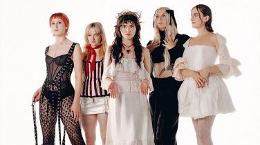 Five women wearing vintage clothes stand in a line. Three of them stare down the lens whilst the other two look into the distance.