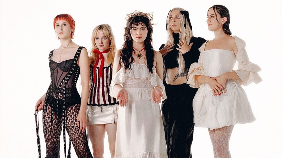 Five women wearing vintage clothes stand in a line. Three of them stare down the lens whilst the other two look into the distance.