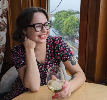 A woman in glasses smiling with a glass of wine.