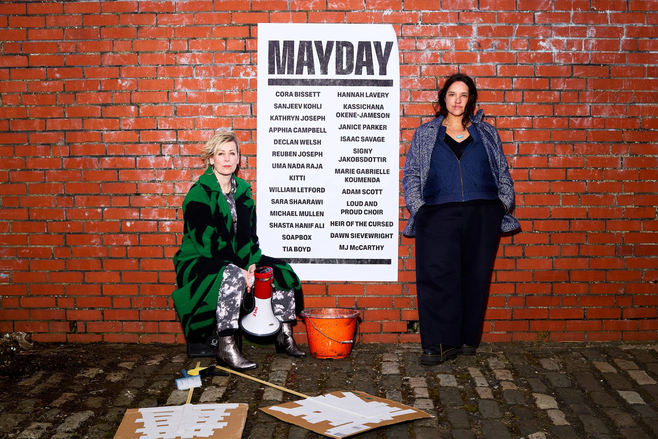 Cora Bissett, seated holding a megaphone, and Hannah Lavery, leaning against a red brick wall featuring a fly poster for Mayday. A pasting bucket and two face-down placards rest on the ground.