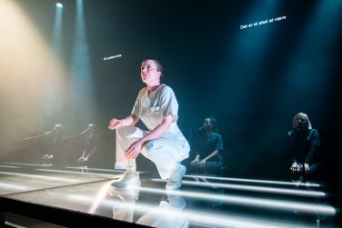 Lærke Schreve Engelbrecht crouching on an illuminated glass stage, with cast members kneeling behind her.