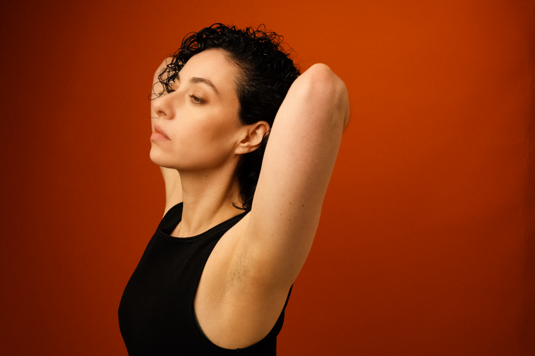 : A profile shot of the artist SHHE wearing a black sleeveless top. Her arms are raised with her hands clasped behind her head, and she is looking off to the left with a calm expression. The background is a warm, solid rust-orange.