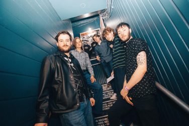 The five members of the band Jutebox posing on a narrow, enclosed staircase with blue wooden walls. The band members are staggered at different heights on the steps. The member in the front left wears a black leather jacket over a graphic tee, while the member in the front right wears a black patterned shirt with his arm tattoos visible. Framed posters are partially visible on the wall behind them.