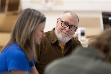 A close-up photograph of actor Barrie Hunter seated at a table during rehearsals. He has a grey beard, wears black-framed glasses and a brown corduroy shirt, and is smiling gently towards the camera. To his left, the side profile of Dawn Steele in a blue t-shirt is softly out of focus.