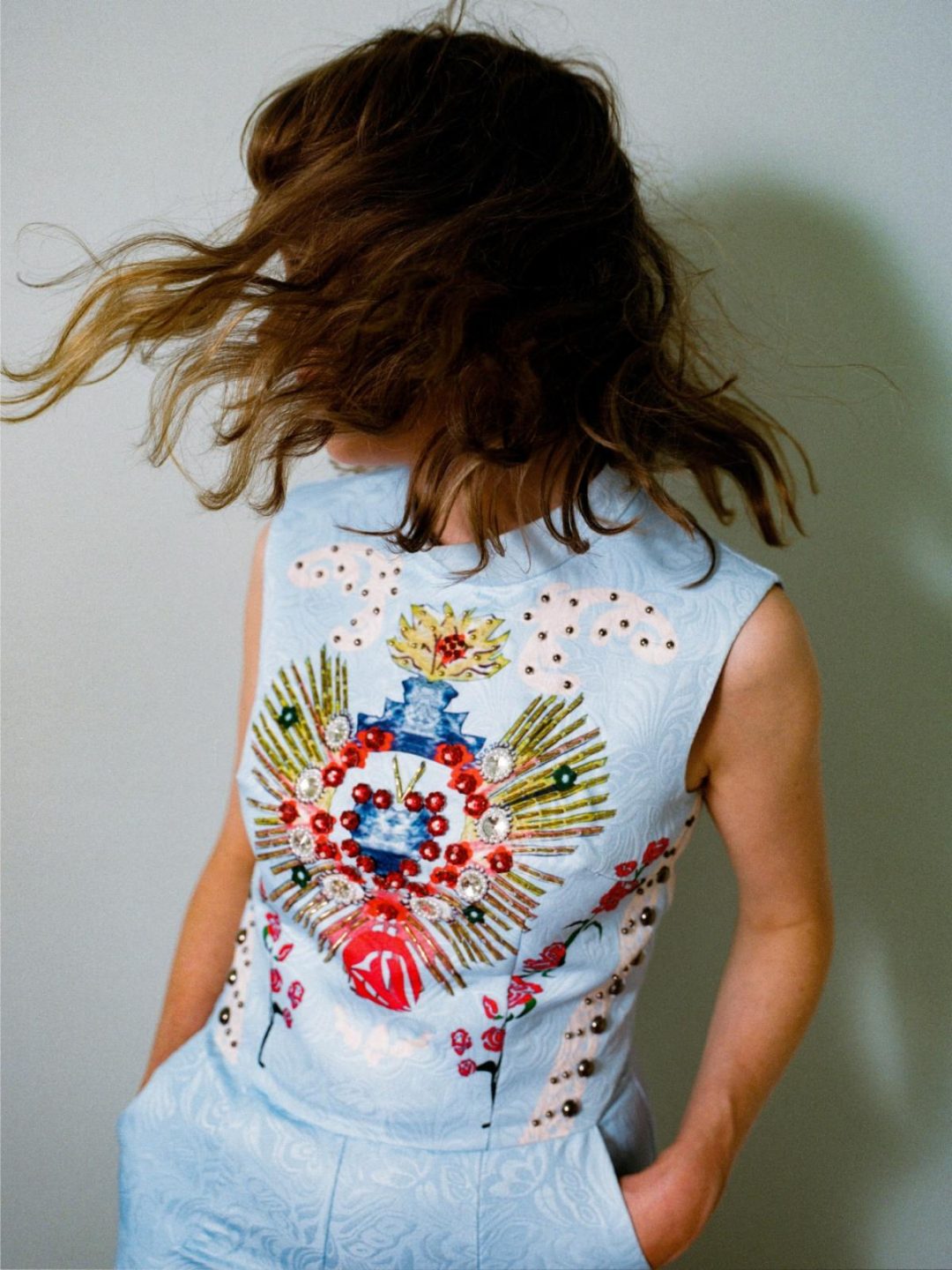 Carla J Easton standing against a plain background, looking away with her hair blowing wildly across her face. She is wearing a light blue sleeveless top with a colorful, ornate embroidered design on the chest.