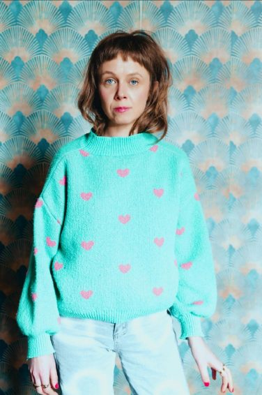 Carla J Easton wearing a light green jumper with pink hearts, standing against blue and gold patterned wallpaper.