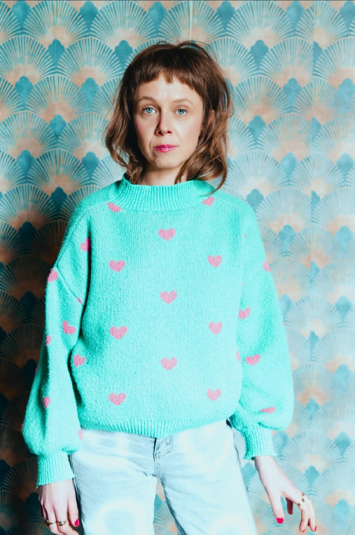 Carla J Easton wearing a light green jumper with pink hearts, standing against blue and gold patterned wallpaper.