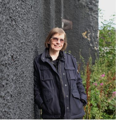 A photograph of Frances Crawford leaning against a heavily textured, dark grey outdoor wall. She has short brown hair, wears glasses, and is smiling directly at the camera. She is wearing a dark blue, multi-pocketed jacket over a dark shirt. To the right of the frame, tall green weeds with small pink flowers are visible growing alongside the wall.