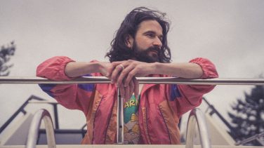 A photograph of Gurry Wurry standing outdoors against an overcast sky with out-of-focus trees in the background. He has long dark hair and a full beard, and is looking thoughtfully off to the right, away from the camera. He is resting his forearms on a silver metal railing. He is wearing a retro-style windbreaker jacket with bright pink, purple, and orange colour-blocked panels over a graphic t-shirt.