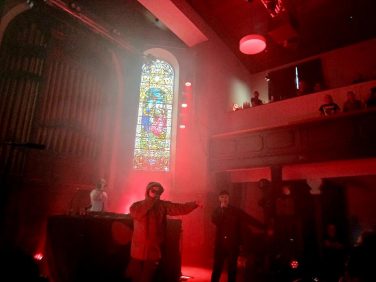 The band Kneecap performing live on stage at St Luke's. The stage is illuminated by intense red lighting and heavy atmospheric smoke. In the background, a large, illuminated stained glass window is flanked by a pipe organ on the left and a balcony with audience members on the right. On stage, a DJ stands behind a desk on the left, while two vocalists are at the front; one in a hooded jacket and sunglasses gestures outwards, and the other stands to the right in dark clothing holding a microphone.