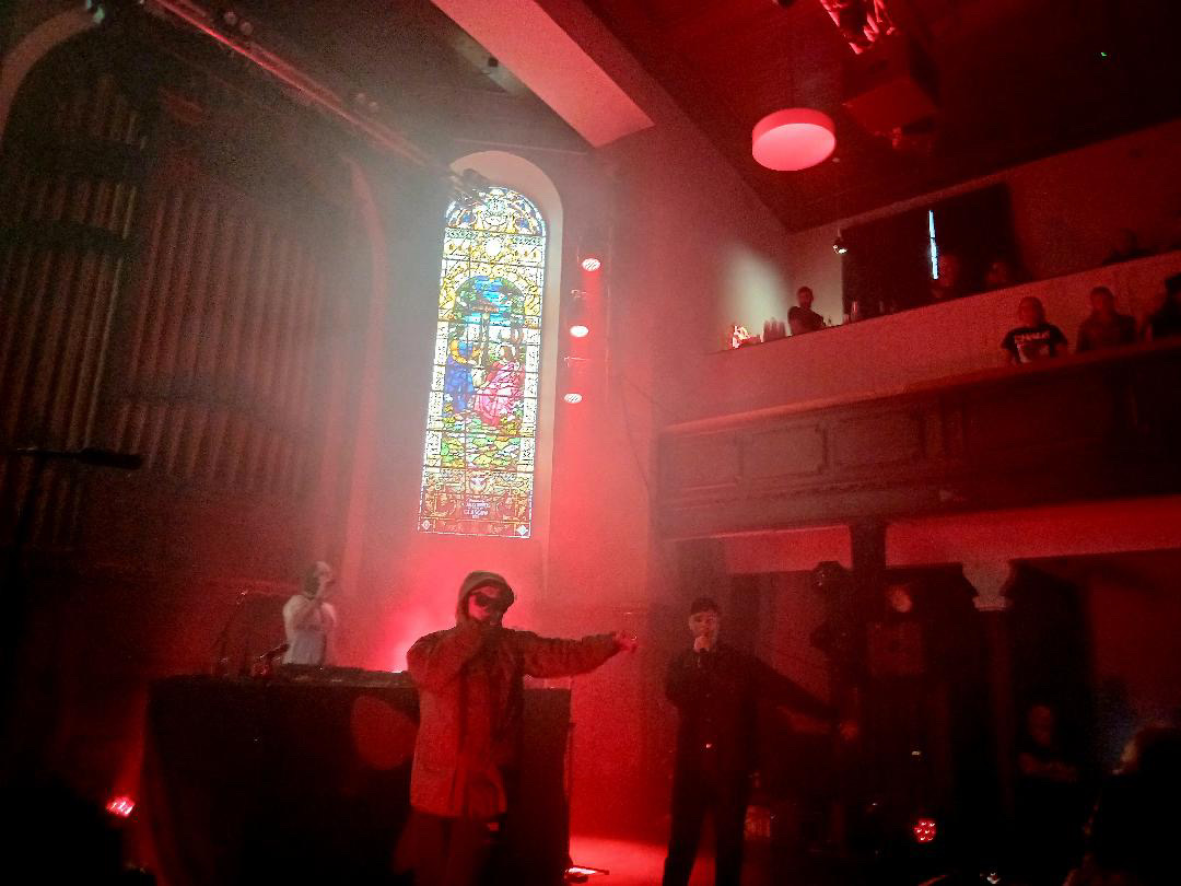 The band Kneecap performing live on stage at St Luke's. The stage is illuminated by intense red lighting and heavy atmospheric smoke. In the background, a large, illuminated stained glass window is flanked by a pipe organ on the left and a balcony with audience members on the right. On stage, a DJ stands behind a desk on the left, while two vocalists are at the front; one in a hooded jacket and sunglasses gestures outwards, and the other stands to the right in dark clothing holding a microphone.
