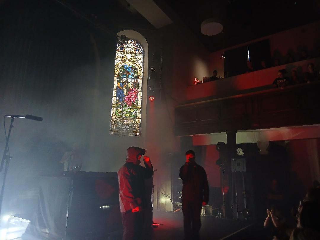 The band Kneecap performing live at St Luke's. The stage is filled with smoke and lit by red spotlights. A large, brightly lit stained glass window is visible directly behind the performers. On stage, a DJ is partially obscured by smoke behind a desk on the left. In the centre, a vocalist in a hooded jacket faces a second vocalist to the right, who is dressed in dark clothing and holding a microphone. Silhouettes of audience members are visible in the foreground and on the balcony to the right.
