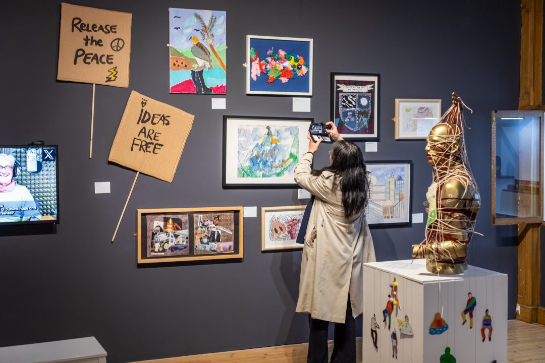 A gallery visitor in a light trench coat takes a photo of an artwork on a dark grey wall filled with various mixed-media pieces, paintings, and cardboard signs.