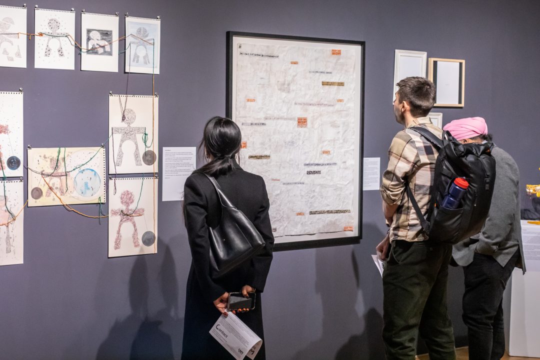 Three people in a gallery, closely examining a large, framed, textured white artwork with small text boxes scattered across it.