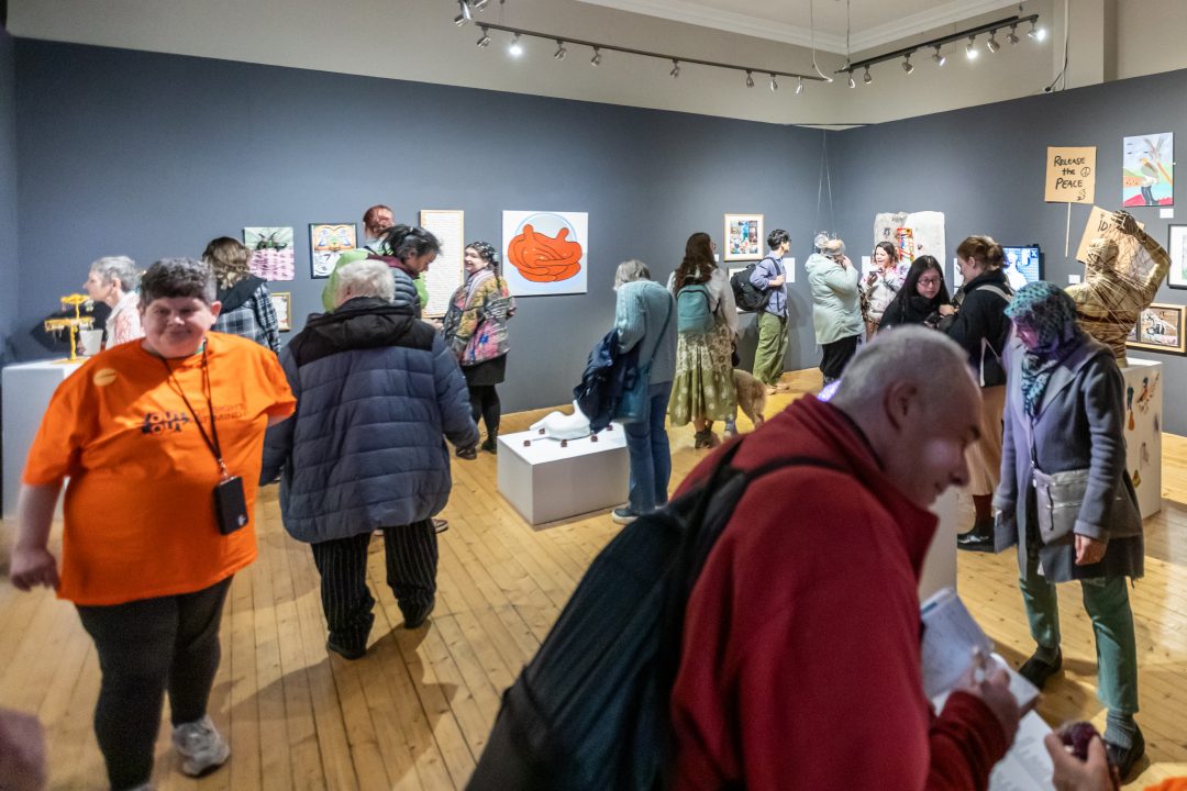 A wide shot of a busy gallery space during an exhibition opening, with numerous attendees walking around and viewing various artworks displayed on dark grey walls and white plinths.