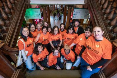 A group photo of the OOSOOM volunteers standing together on a grand wooden staircase, all smiling and wearing matching bright orange "OUT OF SIGHT OUT OF MIND" t-shirts.