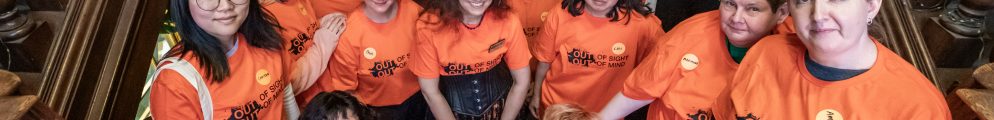 A group photo of the OOSOOM volunteers standing together on a grand wooden staircase, all smiling and wearing matching bright orange "OUT OF SIGHT OUT OF MIND" t-shirts.