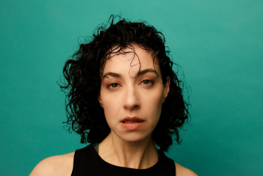 A portrait of SHHE facing forward with a neutral expression. Her dark, wet, curly hair falls loosely around her face, with a few strands resting on her forehead. She is wearing a simple black sleeveless top set against a vibrant green-teal background.
