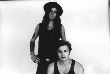 A black and white promotional photo of the band Yacht Club. The standing member has long wavy hair, a wide-brimmed hat, arm tattoos, and wears a dark tank top. He stands with his hands on his hips behind the second member, who is seated in the foreground with short hair and a dark tank top. Both are looking directly at the camera.