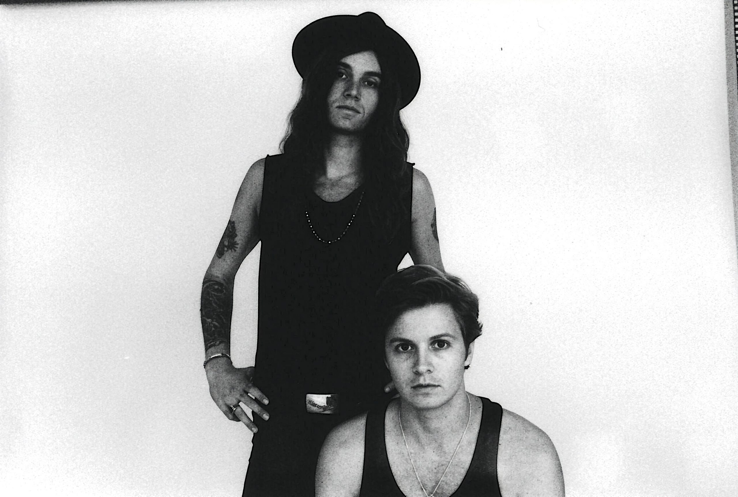 A black and white promotional photo of the band Yacht Club. The standing member has long wavy hair, a wide-brimmed hat, arm tattoos, and wears a dark tank top. He stands with his hands on his hips behind the second member, who is seated in the foreground with short hair and a dark tank top. Both are looking directly at the camera.