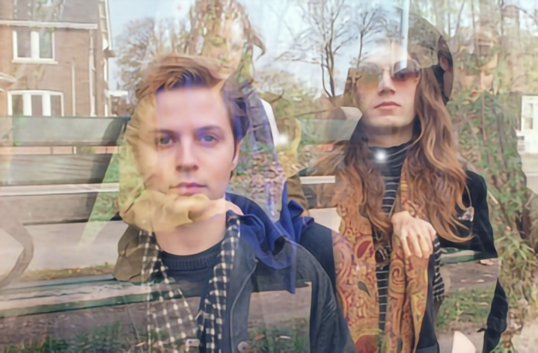 A layered, double-exposure portrait of the band Yacht Club. On the left foreground is the member with short hair, wearing a dark jacket and a checkered scarf. Overlaid on the right is the member with long hair, wearing sunglasses and a colorful paisley scarf. The two figures are semi-transparent, blending into a background scene of a wooden park bench and trees.