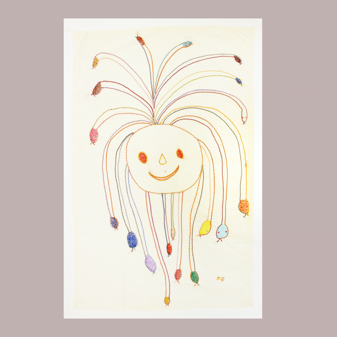 playful cross-stitch embroidery on white fabric depicting a round, smiling face with long, colorful, snake-like tendrils extending outwards from the head, ending in distinct snake-like shapes.