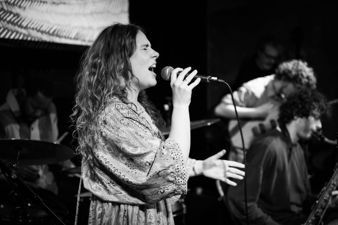Ellyn Oliver singing on a stage with bandmates behind her.