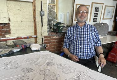 A photograph of artist James Gladwell, a smiling man with a beard and glasses wearing a blue and white checkered shirt, sitting at a work table in a studio with a piece of fabric featuring an intricate line-drawn embroidery pattern in front of him.