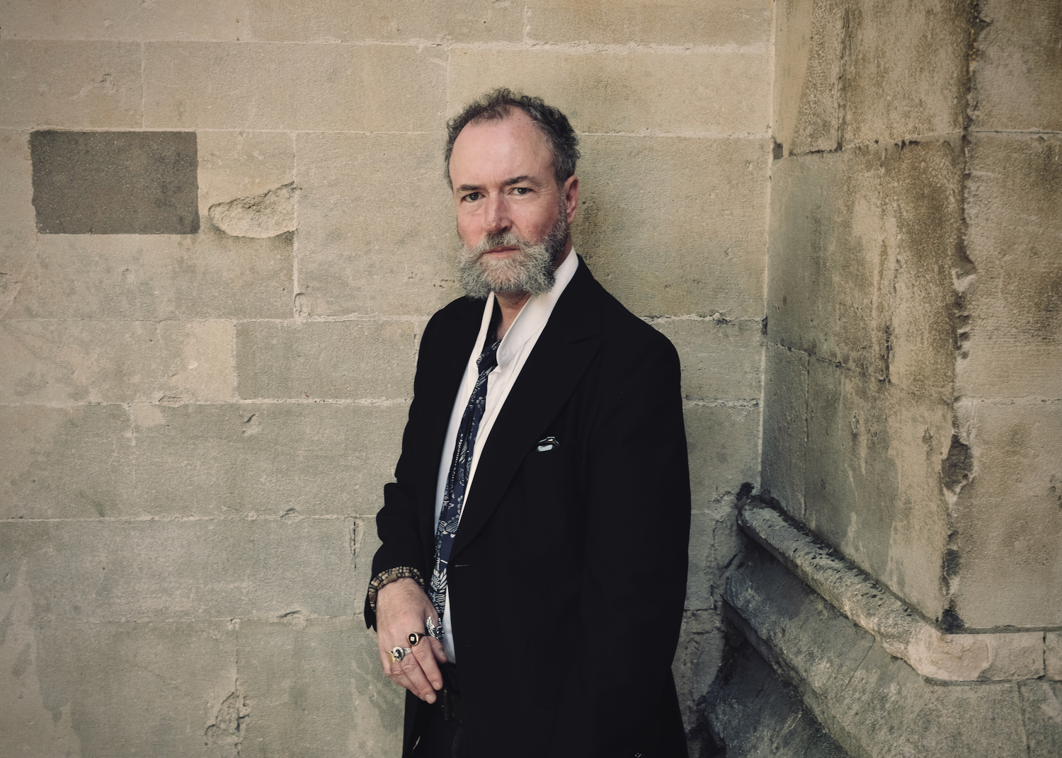 A photo of author David Keenan, who has a grey beard and is wearing a dark suit with a patterned tie, standing against a stone wall.