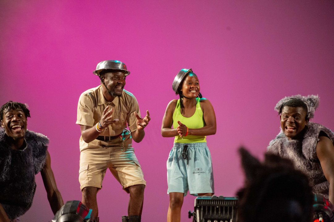 A scene from the stage production Afronauts featuring four performers against a bright pink backdrop. A man in a beige utility outfit and a woman in a neon green tank top and light blue shorts both wear silver metal bowls on their heads as makeshift helmets, while performers on either side are dressed in dark, fuzzy, animal-like costumes.