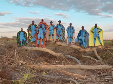 A group of nine performers in matching light blue jumpsuits and vibrant, patterned superhero-style capes posing triumphantly in a row on a large, fallen tree trunk. The scene is set outdoors in a rural landscape under a soft sky.