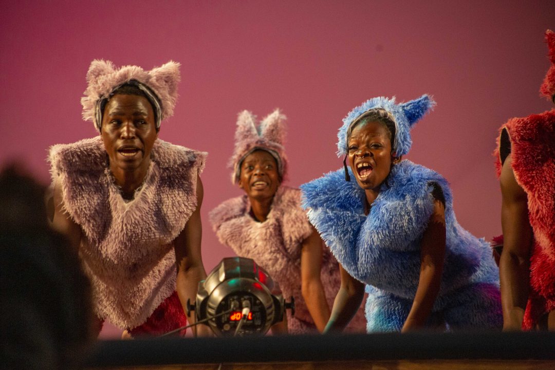 A scene from the stage production Afronauts featuring four performers against a bright pink backdrop. A man in a beige utility outfit and a woman in a neon green tank top and light blue shorts both wear silver metal bowls on their heads as makeshift helmets, while performers on either side are dressed in dark, fuzzy, animal-like costumes.