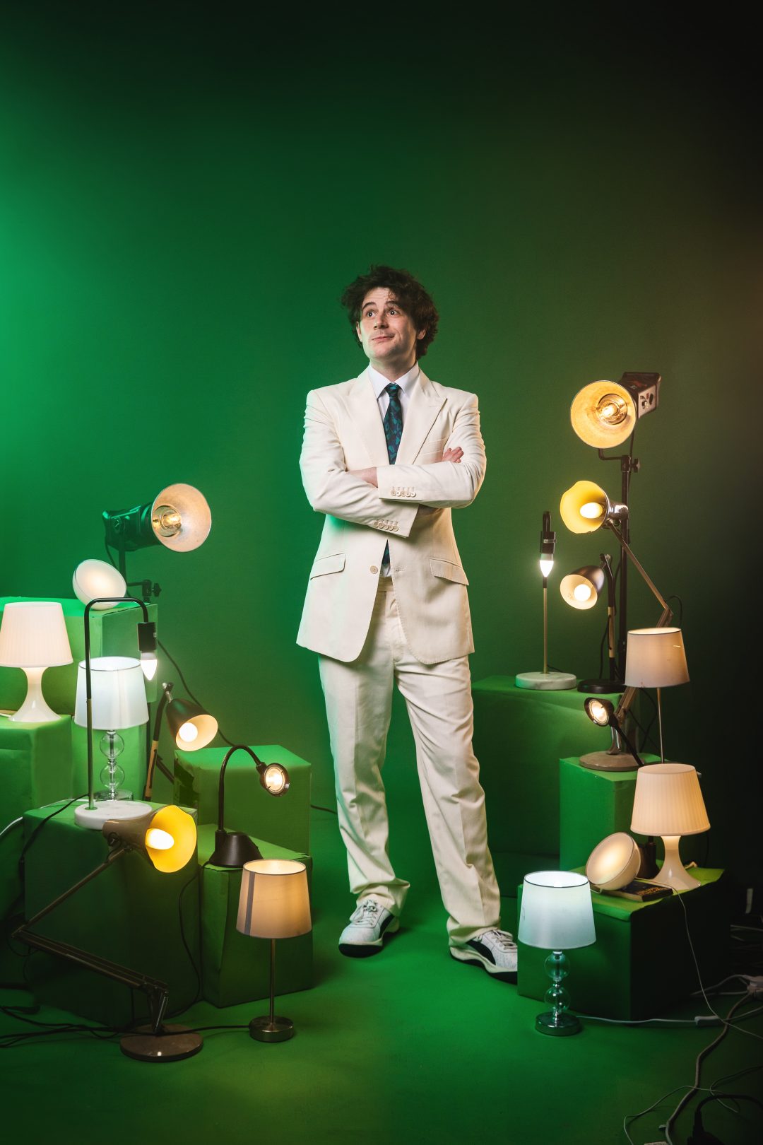 A full-body portrait of comedian Alex Kitson, dressed in a cream suit and trainers, standing with arms crossed and looking up. He is surrounded by numerous illuminated table and floor lamps of different styles, placed on green blocks against a solid green studio background.