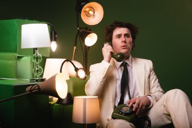 Comedian Alex Kitson, wearing his cream suit, sits among the lamps on green blocks against the green background. He holds a vintage green rotary telephone to his ear, looking concerned