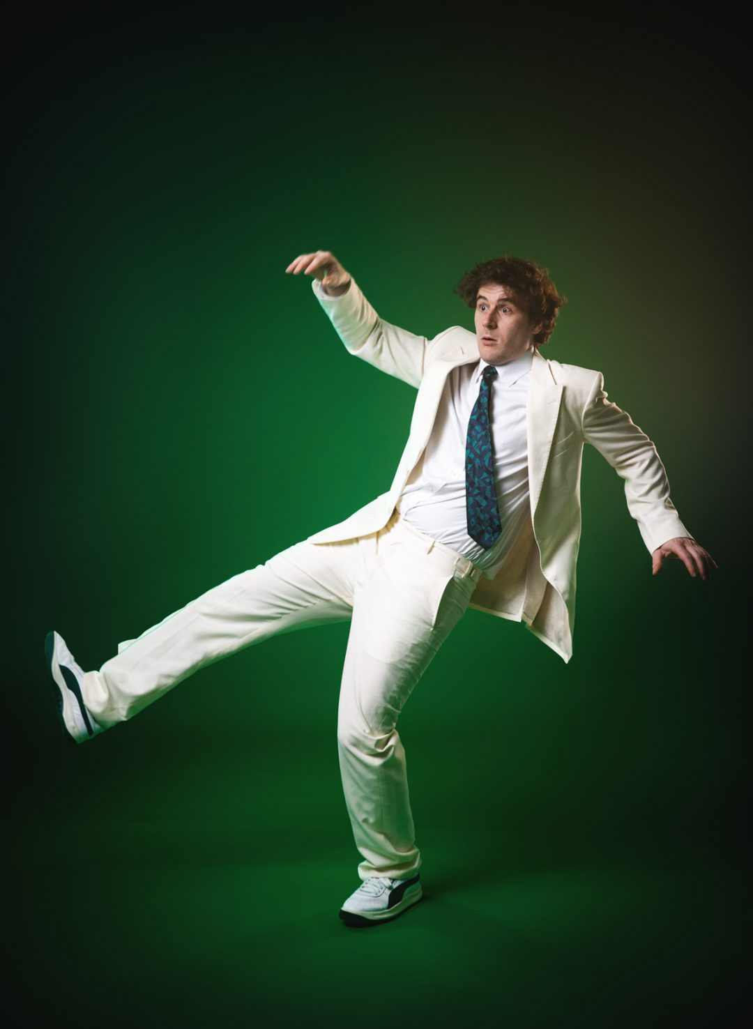 Comedian Alex Kitson, in his cream suit, is captured mid-action on the green studio floor. He leans back sharply, kicking one leg into the air with a surprised expression, as if reacting to an unseen event.