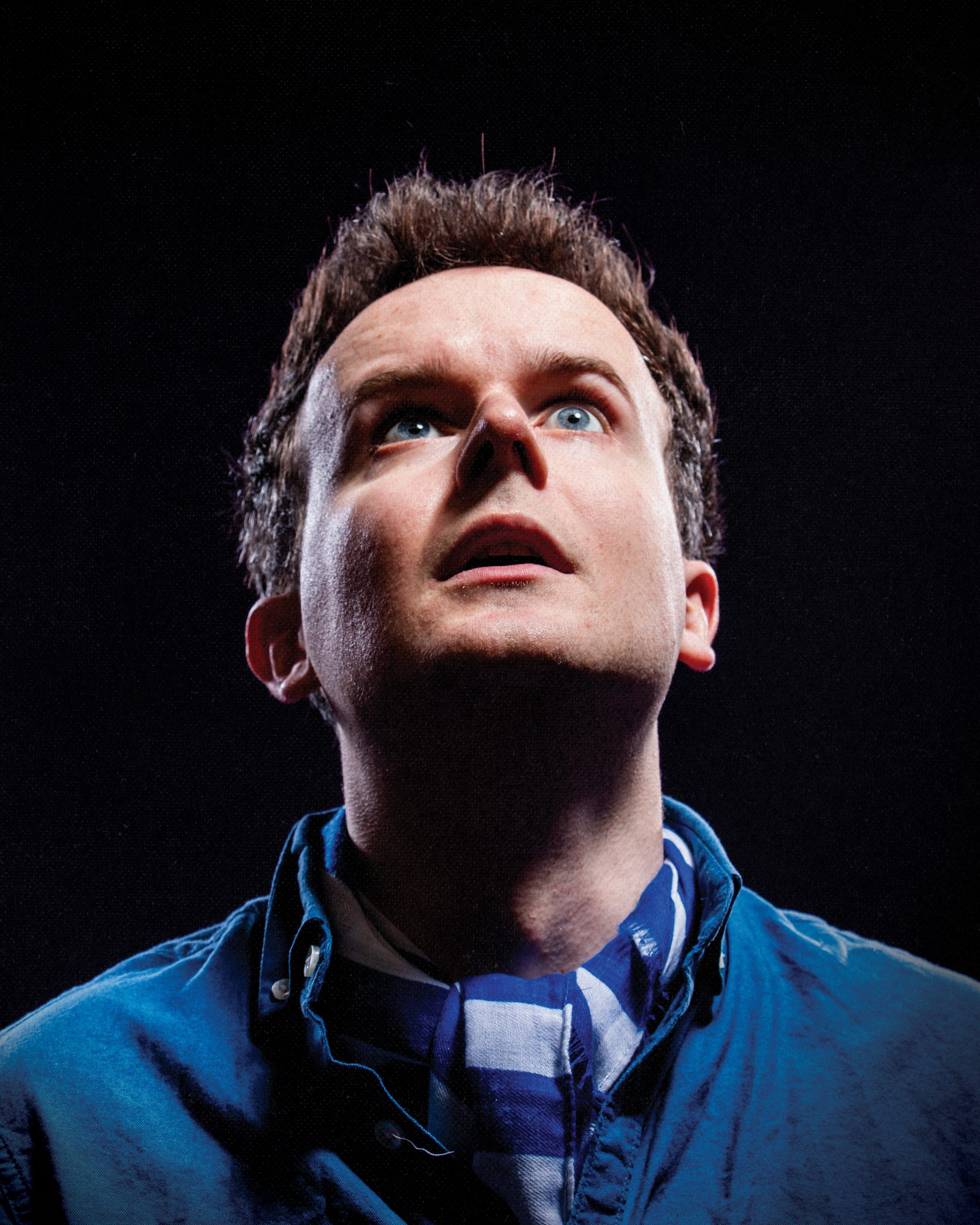Andrew Doherty looking upward with a startled expression against a dark background, wearing a blue shirt and patterned scarf.