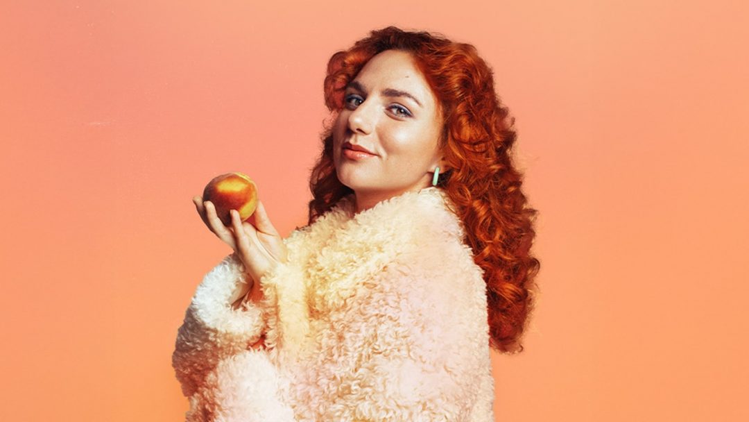 Comedian Ania Magliano holding a peach in a fluffy coat against a peach-coloured background for her show Peach Fuzz.