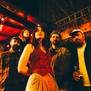 Bikini Body band members posing together under warm red lighting outdoors at night.