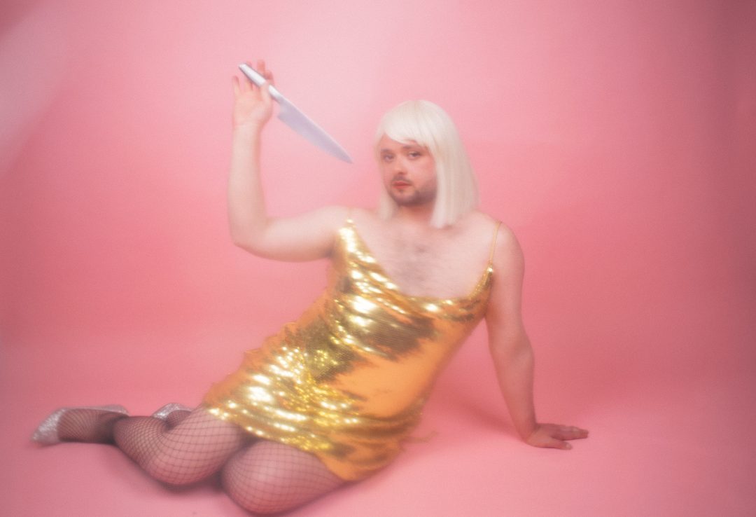 Performer Craig Manson in a blonde bob wig and gold sequined dress, sitting on a pink floor and holding a large knife.