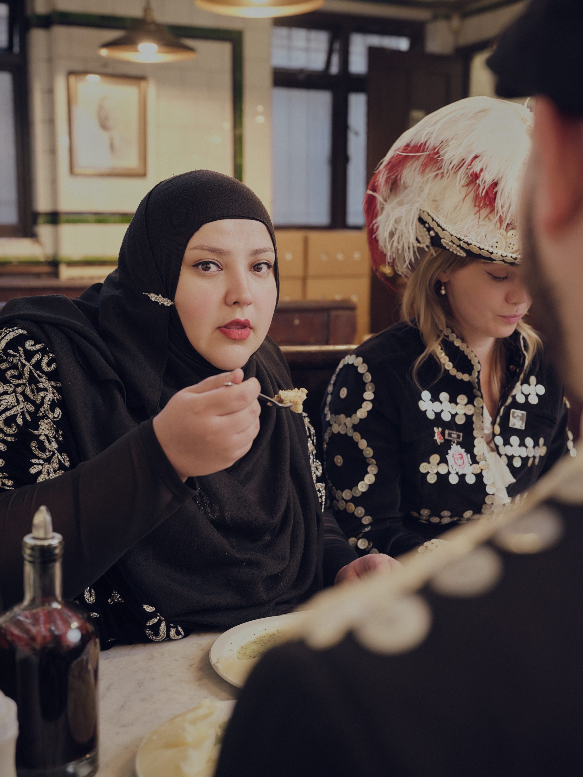 A woman in a black hijab and embroidered dress eats at a cafe table with a man in the foreground. Beside her, a woman wears an elaborate feathered military hat and jacket. The background features a vintage tiled cafe interior.