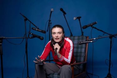 Olivia Caw sitting in a plaid armchair holding a microphone against a blue background surrounded by multiple microphone stands for the Edinburgh Fringe theatre production Common Tongue.